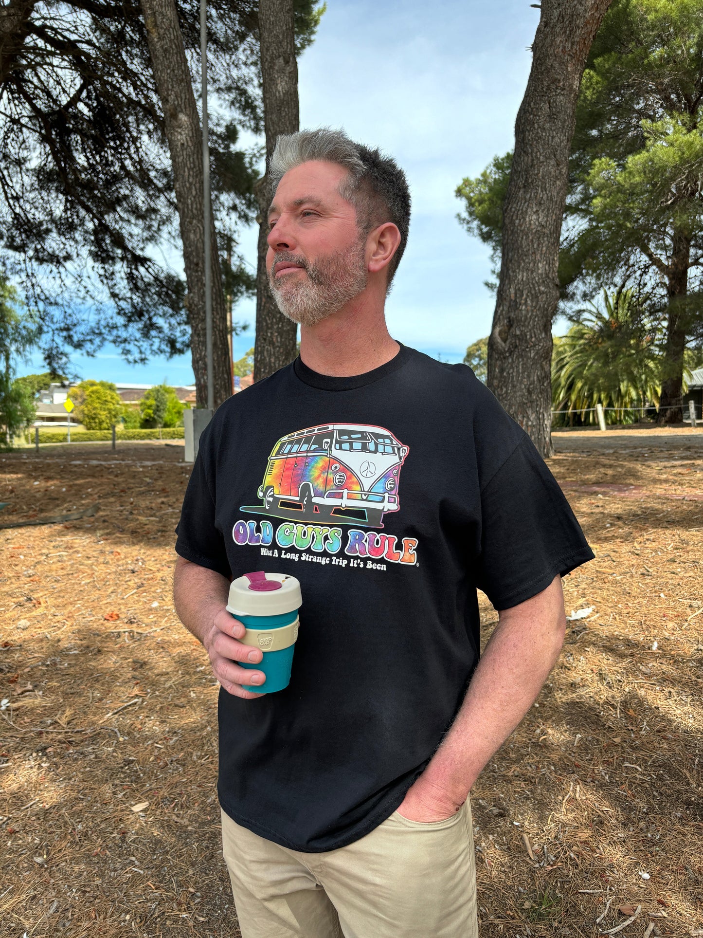 Matt from The Bloke Shop is wearing Old Guys Rule Trippin T Shirt. Rainbow Kombi van logo and text reads: what a long strange trip it's been.  He's holding a coffee cup and standing in the woods with a wry look on his face.