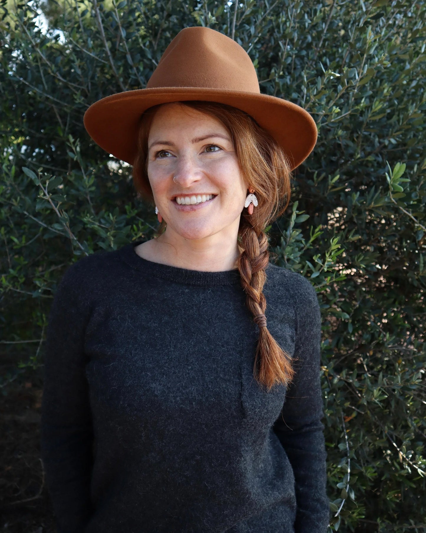 Best Ladies Akubra Hats. Woman wearing an Akubra Traveller Hat in Rust. She is smiling and looking far off, there is Australian shrubs behind her.